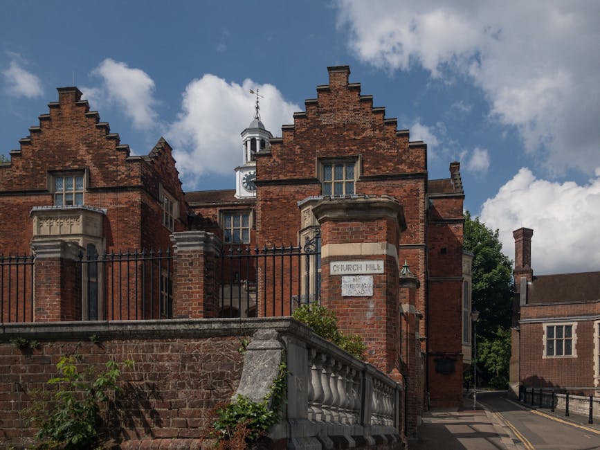 A historic red brick building with a stepped gable roof, decorative stone window surrounds, and a clock tower with a weather vane on top, situated behind a wrought iron fence mounted on a brick wall. 