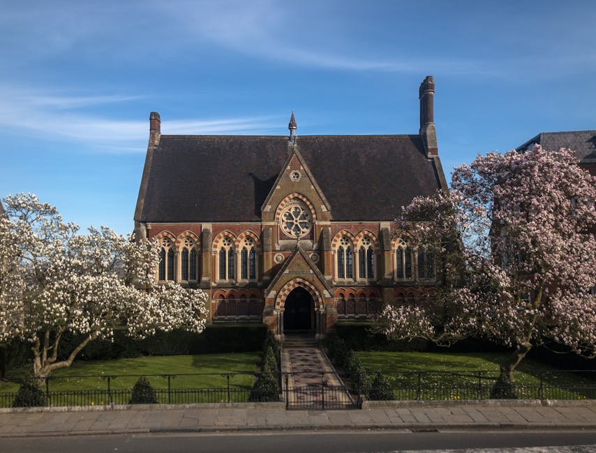 A historic red brick building with a stepped gable roof, decorative stone window surrounds, and a clock tower with a weather vane on top, situated behind a wrought iron fence mounted on a brick wall. 