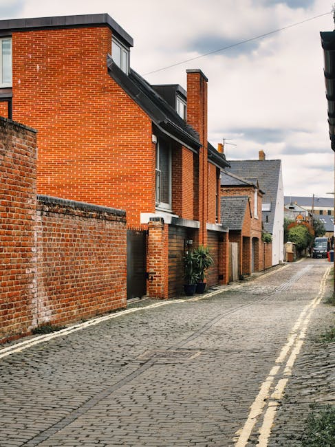 A narrow residential alleyway with a paved stone pathway leading uphill between two brick walls. The left wall, painted white, features small windows and a hidden doorway or window opening, while the 