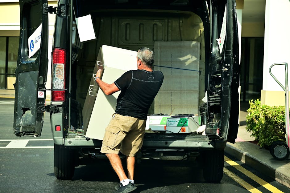 A man wearing a black t-shirt, beige cargo shorts, and sports shoes is loading a white cabinet into the back of a black moving van parked on a paved street. The van's rear doors are open, revealing a 